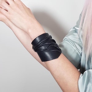 Puede incluir: Una pulsera brazalete ancha de cuero negro que presenta una serie de pliegues horizontales y fruncidos que envuelven la mu&ntilde;eca. El accesorio tiene un acabado mate liso y un dise&ntilde;o estructurado que cubre una parte del antebrazo.