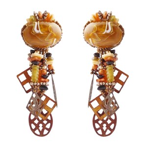 May include: A pair of ornate earrings with a warm color palette. Each earring features a large, oval-shaped amber-colored stone with white swirls, surrounded by a gold-toned metal frame. Below the stone hang various beads and geometric shapes.