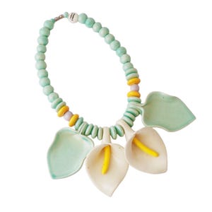 May include: A ceramic necklace featuring a light blue beaded strand with yellow and pink accents. The necklace has three white calla lily pendants with yellow stamens and light green leaves. The clasp has the text "Opaque 2 Oceans".