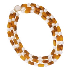Puede incluir: Collar de tres hilos con filas alternas de cuentas redondas blancas y cuentas rectangulares de color ámbar. El collar se asegura con un cierre dorado. El diseño es clásico y elegante, adecuado para diversas ocasiones.