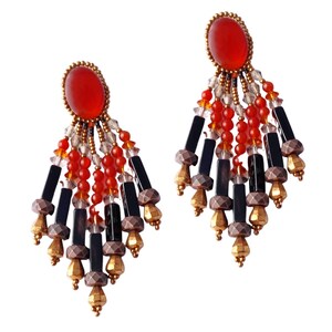 May include: A pair of statement earrings featuring a large oval red stone at the top, surrounded by gold-colored beads. Below, the earrings have strands of red beads, black rectangular beads, and gold-colored geometric shapes, creating a layered, dangling effect.