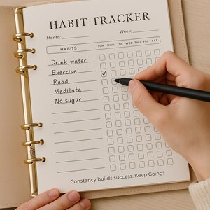 Puede incluir: Un cuaderno beige para el seguimiento de h&aacute;bitos con anillos de metal dorado. El cuaderno est&aacute; abierto, mostrando una tabla de seguimiento de h&aacute;bitos con el t&iacute;tulo "HABIT TRACKER". La tabla incluye una lista de h&aacute;bitos y una cuadr&iacute;cula para el seguimiento diario. Una mano sostiene un bol&iacute;grafo negro, marcando una casilla.