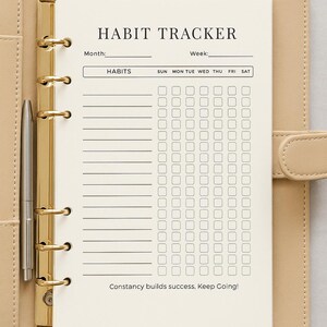 Puede incluir: Un cuaderno beige para el seguimiento de h&aacute;bitos con anillos dorados y un bol&iacute;grafo. El cuaderno est&aacute; abierto en una p&aacute;gina que dice "HABIT TRACKER" y tiene espacios para rastrear h&aacute;bitos. El texto "Constancy builds success. Keep Going!" est&aacute; en la parte inferior.