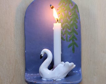 Può includere: Un candelabro da parete in ceramica con una piastra posteriore ad arco viola decorata con un motivo ad albero dipinto in verde. Una statuetta di cigno bianco funge da base per una candela bianca, accesa e con una fiamma gialla.