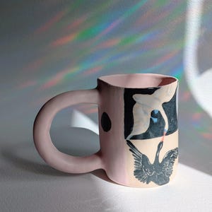 May include: A pink ceramic mug with a large, rounded handle. The mug features a black and white illustration of a swan. A black dot is on the side of the mug. The mug is on a white surface.