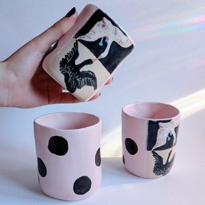 Puede incluir: Tazas de cerámica rosa con lunares negros e ilustraciones de cisnes. Una taza se sostiene, mostrando un diseño de cisne en blanco y negro. Las otras dos tazas están sobre una superficie blanca, con interior rosa.