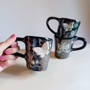 May include: Three ceramic mugs with butterfly designs. The mugs are black with iridescent glaze and feature white butterfly accents. Two of the mugs are stacked, while the third mug is held in a hand.