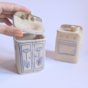 Könnte beinhalten: Zwei Keramikbehälter, einer in Form eines Waschbeckens mit Deckel und der andere in Form eines Herds. Der Waschbeckenbehälter ist offen und gibt sein Inneres frei, mit blauen Blumendesigns. Der Herd hat eine blaue Ofentür und Brennerdetails.