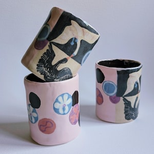 May include: Three cylindrical ceramic cups with hand-painted designs. One cup features black bird illustrations on a beige background. Another cup is light pink with blue, purple, and orange circular floral motifs. The third cup combines these elements.