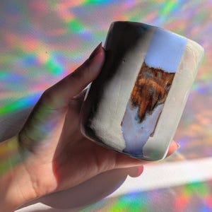 Può includere: Una tazza cilindrica in ceramica con finitura esterna grigio chiaro opaco. Una fascia verticale di smalto blu chiaro lucido corre lungo il centro, caratterizzata da una zona testurizzata marrone scuro con toni metallici al centro del blu.