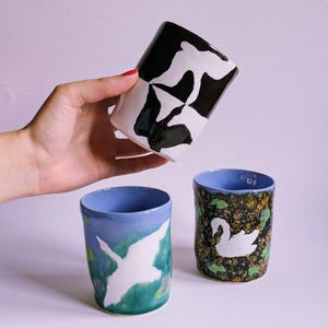 Puede incluir: Tres tazas de cerámica con diseños distintos. Una taza presenta un patrón abstracto en blanco y negro. Otra taza muestra la silueta de un ave blanca sobre un fondo azul y verde. La tercera taza muestra un cisne blanco sobre una superficie moteada.