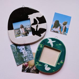 May include: Two handmade ceramic picture frames with irregular shapes. One frame is black and white with a swan motif, holding a photo of a person. The other is green with white swan and lily pad designs. Three loose square photographs are included.