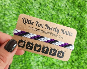 May include: A black, white, and purple friendship bracelet with a brown card that says "Little Fox Nerdy Knits" and includes a website address and social media icons.