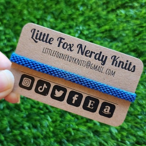 May include: A brown card with the text "Little Fox Nerdy Knits" and the email address "littlefoxnerdyknits@gmail.com". The card has a blue cord wrapped around it and icons for Instagram, TikTok, Twitter, Facebook, Etsy, and Amazon.