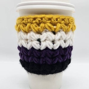 Puede incluir: Una taza de café desechable blanca con tapa blanca, envuelta en un protector de ganchillo. El protector presenta rayas horizontales en amarillo, blanco, morado y negro. El protector está diseñado para aislar la taza y proteger las manos.