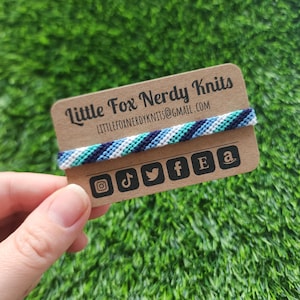 May include: A brown card with the text "Little Fox Nerdy Knits" and the email address "littlefoxnerdyknits@gmail.com". A blue, white and green friendship bracelet is wrapped around the card. Social media icons for Instagram, TikTok, Twitter, Facebook, Etsy and Amazon are printed on the card.