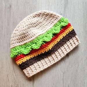 Puede incluir: Un gorro de crochet con forma de hamburguesa. El gorro es beige con detalles de lechuga verde, tomate rojo, queso amarillo y carne marrón.