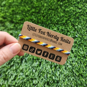 May include: A brown card with the text "Little Fox Nerdy Knits" and the email address "littlefoxnerdyknits@gmail.com". A colorful friendship bracelet is wrapped around the card. Social media icons are printed on the card.