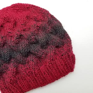 Op de afbeelding: Een rode en zwarte gebreide beanie met een kabelbreipatroon.