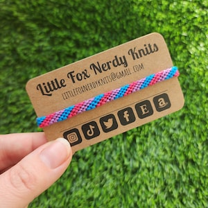 May include: A brown card with the text "Little Fox Nerdy Knits" and the email address "littlefoxnerdyknits@gmail.com". A blue, pink, and purple friendship bracelet is wrapped around the card. Social media icons for Instagram, TikTok, Twitter, Facebook, Etsy, and Amazon are printed on the card.
