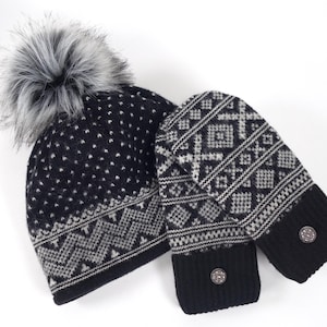 May include: A black and grey knit beanie with a faux fur pom-pom and a matching pair of mittens. Both items feature a geometric pattern, a solid black ribbed cuff, and a small circular metallic button detail on the wrist of each mitten.