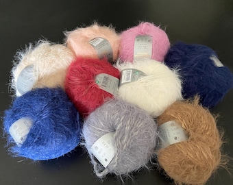 Puede incluir: Una colección de diez ovillos de lana difusos en varios colores, incluyendo melocotón, rosa, azul oscuro, crema, rojo, blanco, azul marino, gris y bronceado. Cada ovillo tiene una etiqueta de papel con instrucciones de cuidado y texto.