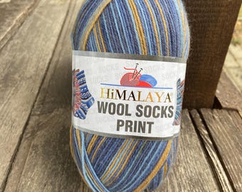 May include: A ball of Himalaya Wool Socks Print yarn in shades of blue, gold, and navy. The yarn is wrapped with a label that reads "Himalaya Wool Socks Print". The yarn is sitting on a wooden surface.