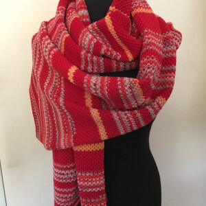 Könnte beinhalten: Ein gestrickter Schal in Rot, Orange und Grau mit Streifen. Der Schal ist über eine schwarze Schaufensterpuppe gelegt.