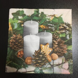 May include: Three white candles with a gingerbread cookie and pine cones on a green and brown background.