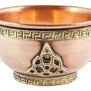 May include: A copper-colored bowl with a decorative gold-colored trim and a Celtic knot design. The bowl has a rounded shape with a wide opening and a small base. The interior of the bowl has a shiny, reflective surface.