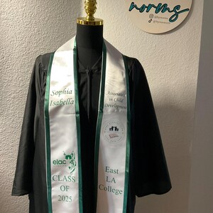 May include: A white graduation sash with green trim. The sash has the text "Sophia Isabella" on one side and "Associates in Child Development" on the other side. The sash also has the text "elac" and "East LA College" on it. The sash is worn by a person in a black graduation gown.