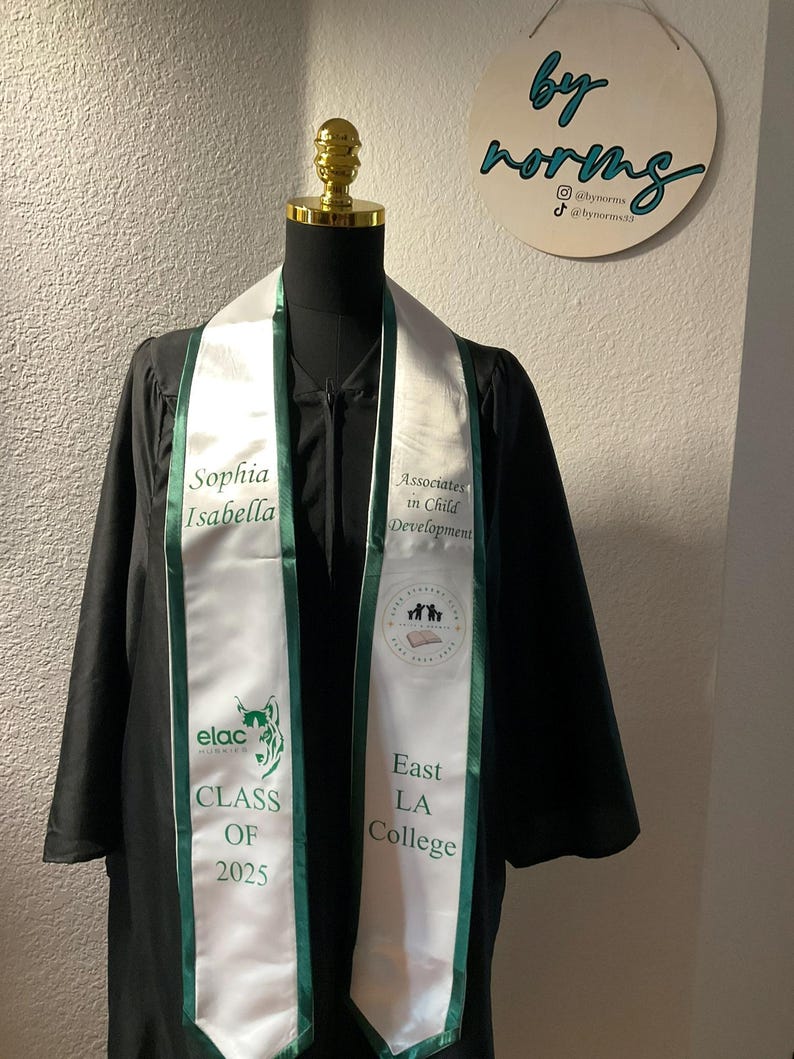 May include: A white graduation sash with green trim. The sash has the text "Sophia Isabella" on one side and "Associates in Child Development" on the other side. The sash also has the text "elac" and "East LA College" on it. The sash is worn by a person in a black graduation gown.