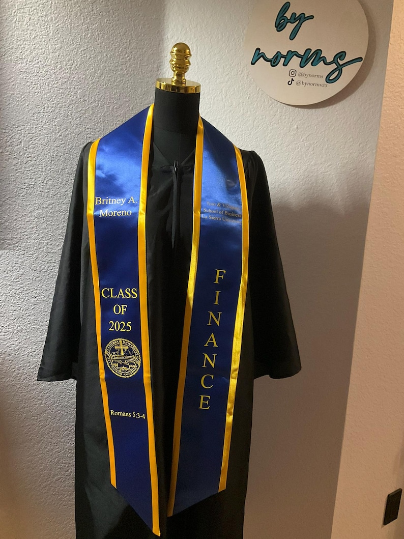 May include: A blue and gold graduation sash with the text "Britney A. Moreno", "Class of 2025", "Romans 5:3-4", and "Finance".