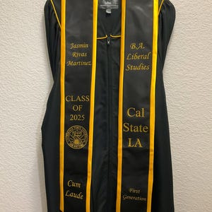 May include: Black graduation stole with gold trim and embroidered text. The text reads "Jasmin Rivas Martinez", "B.A. Liberal Studies", "Class of 2025", "Cal State LA", "Cum Laude", and "First Generation".