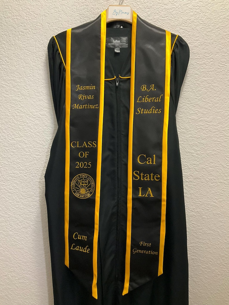 May include: Black graduation stole with gold trim and embroidered text. The text reads "Jasmin Rivas Martinez", "B.A. Liberal Studies", "Class of 2025", "Cal State LA", "Cum Laude", and "First Generation".