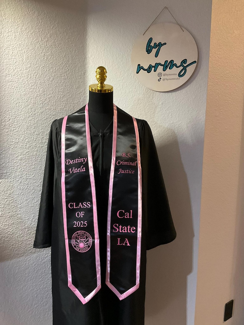 May include: Black graduation gown with a pink sash. The sash has the text "Destiny Vitela", "Class of 2025", "Cal State LA", and "B.S. Criminal Justice" embroidered on it.