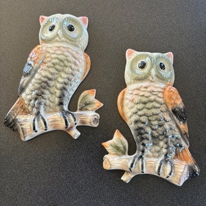 May include: Two ceramic owl wall decorations. The owls are perched on a branch with leaves. The owls have large, round eyes, and are painted in shades of grey, brown, and orange. The background is a dark grey.