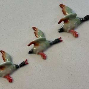 May include: Three ceramic duck figurines painted in green, brown, and white. The ducks are in flight with their wings outstretched.