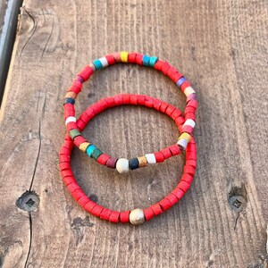 May include: Two red beaded bracelets, one with colorful beads and one with a solid red color. Both bracelets have a wooden bead accent.