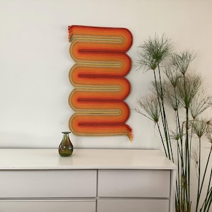 May include: A wall hanging with a wavy, vertical design in shades of orange, red, and tan. The artwork is mounted on a white wall. A small, green glass vase sits on a white dresser below the artwork. Tall green plants are to the right.