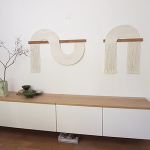 May include: Two white macrame wall hangings with wooden dowels, one in a curved shape and the other in a half-circle shape, hanging on a white wall above a white cabinet with three drawers.