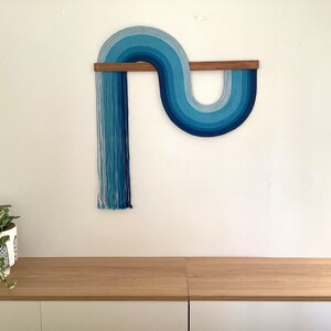 Puede incluir: Un tapiz de pared azul y blanco con un dise&ntilde;o curvo, hecho de hilo, colgado de una barra de madera.