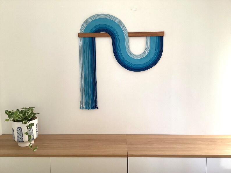 Puede incluir: Un tapiz de pared azul y blanco con un dise&ntilde;o curvo, hecho de hilo, colgado de una barra de madera.