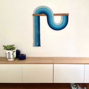 Puede incluir: Un tapiz de pared azul y blanco con un dise&ntilde;o curvo, montado en una barra de madera. El tapiz de pared est&aacute; hecho de hilo y tiene un aspecto moderno y abstracto.