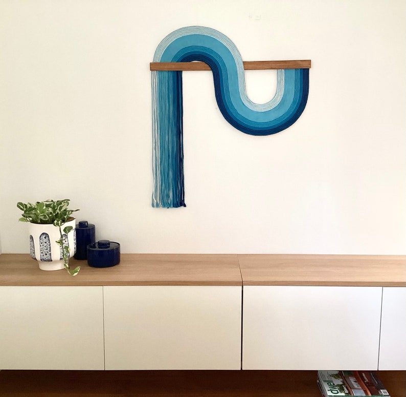 Puede incluir: Un tapiz de pared azul y blanco con un dise&ntilde;o curvo, montado en una barra de madera. El tapiz de pared est&aacute; hecho de hilo y tiene un aspecto moderno y abstracto.