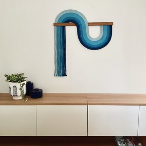 Puede incluir: Una pared colgante azul y blanca con un dise&ntilde;o curvo. La pared colgante est&aacute; hecha de hilo y est&aacute; unida a una varilla de madera.