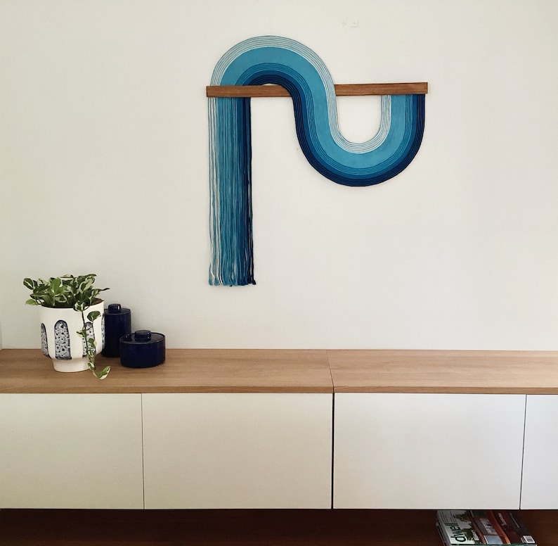 Puede incluir: Una pared colgante azul y blanca con un dise&ntilde;o curvo. La pared colgante est&aacute; hecha de hilo y est&aacute; unida a una varilla de madera.