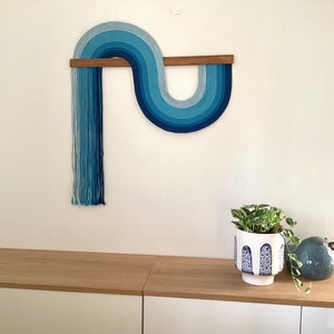 Puede incluir: Una pared colgante de hilo azul y blanco en forma de ola, con una espiga de madera en la parte superior. La pared colgante est&aacute; montada en una pared blanca, y hay una planta en maceta y una figurita de p&aacute;jaro de cer&aacute;mica en un estante de madera debajo.