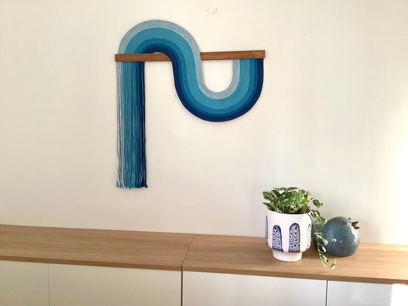 Puede incluir: Una pared colgante de hilo azul y blanco en forma de ola, con una espiga de madera en la parte superior. La pared colgante est&aacute; montada en una pared blanca, y hay una planta en maceta y una figurita de p&aacute;jaro de cer&aacute;mica en un estante de madera debajo.