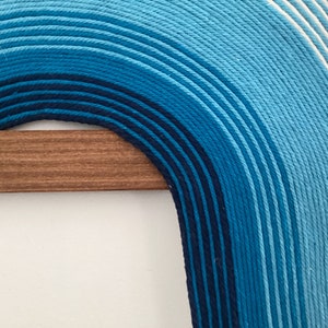 Puede incluir: Una gran pared colgante de hilo azul y blanco en forma de arco. El hilo est&aacute; tejido en un patr&oacute;n degradado, comenzando con azul oscuro en la parte inferior y desvaneci&eacute;ndose a blanco en la parte superior. La pared colgante est&aacute; unida a una varilla de madera.